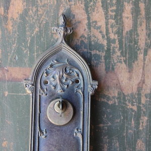 May include: Ornate antique door hardware with a keyhole and decorative floral design. The metal has a weathered, aged appearance with a dark bronze finish. The hardware is mounted on a distressed wooden door.