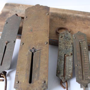 May include: Four antique brass and metal spring scales of varying sizes, arranged on a weathered wooden board. The scales have visible measurement markings and hanging hooks. Some scales have text, including "Excelsior Improved Spring Balance" and "W. & PARTEL".