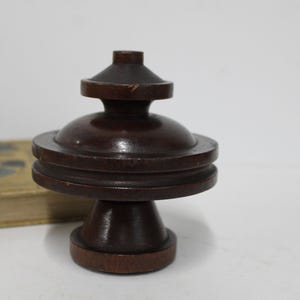 May include: A dark brown, turned wooden object with a tiered design. The object has a rounded top, a central section with two stacked rings, and a flared base. The wood grain is visible. A book is partially visible in the background.