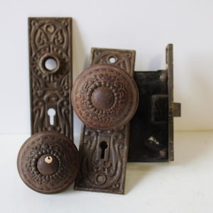 May include: Antique door hardware set, featuring two ornate doorknobs, two backplates, and a mortise lock. The hardware is made of cast iron with a dark brown, aged finish. The doorknobs have a detailed, raised pattern.