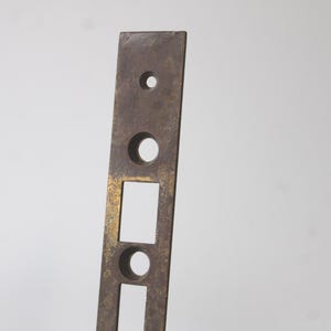 May include: A rectangular, antique metal door strike plate with a dark, aged finish. It features three circular holes and two rectangular cutouts, showing signs of wear and age. The metal has a slightly tarnished, bronze-like appearance.