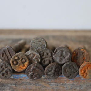 May include: A collection of small, round, antique-style buttons with various embossed designs and numbers. The buttons are primarily dark gray with hints of orange and brown, resting on a weathered wooden surface.