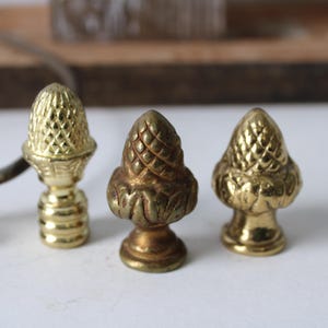 May include: Three brass acorn finials in varying shades of gold. The finials have detailed textures and are displayed on a white surface. One finial is attached to a metal fixture. The acorn-shaped design is a classic decorative element.