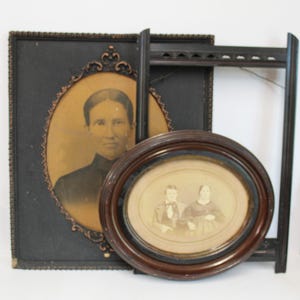 May include: A collection of antique picture frames, including a large black frame with an oval portrait, a dark wooden frame, and a smaller oval frame with a sepia-toned portrait. The frames are of various sizes and styles.