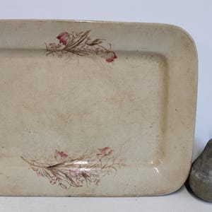 May include: A rectangular off-white ceramic serving platter featuring a delicate brown and pink floral botanical illustration at the top and bottom center, accompanied by a gray pear-shaped decorative object.