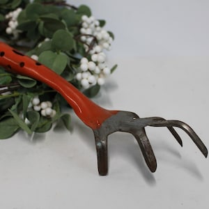 May include: A small garden hand rake with a bright orange handle featuring black dots. The metal tines are curved and aged. The tool is designed for gardening and is set against a backdrop of greenery and white berries.