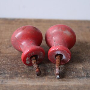 Puede incluir: Dos pomos de madera roja vintage con pintura envejecida y desgastada. Cada pomo tiene una parte superior redondeada y una base cil&iacute;ndrica, con un tornillo de metal oxidado que sobresale de la parte inferior. Los pomos son probablemente para un armario o un caj&oacute;n.