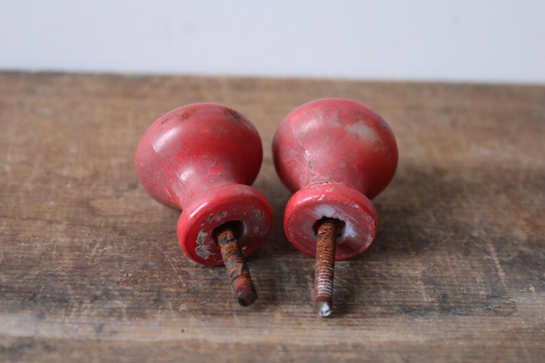 Puede incluir: Dos pomos de madera roja vintage con pintura envejecida y desgastada. Cada pomo tiene una parte superior redondeada y una base cil&iacute;ndrica, con un tornillo de metal oxidado que sobresale de la parte inferior. Los pomos son probablemente para un armario o un caj&oacute;n.
