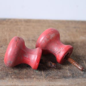 Puede incluir: Dos tiradores de armario rojos vintage con un acabado envejecido. Cada tirador tiene una parte superior redondeada y una base cil&iacute;ndrica, fijada a un tornillo de metal oxidado. Los tiradores est&aacute;n sobre una superficie de madera desgastada.