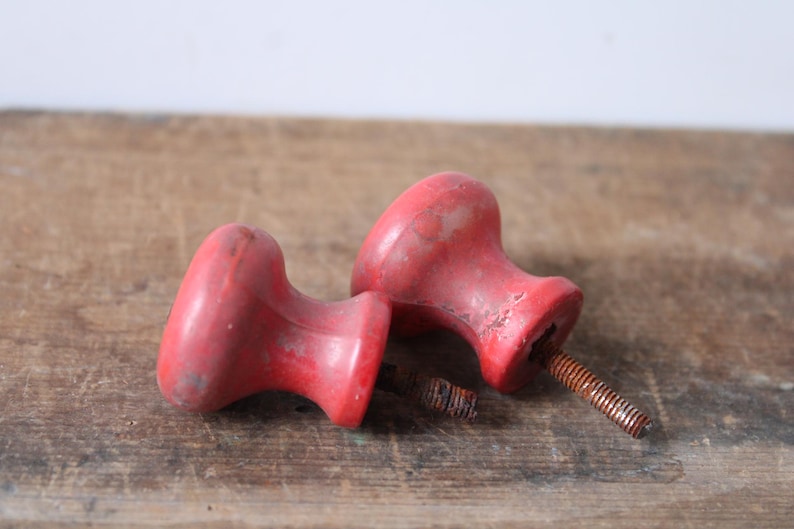 Puede incluir: Dos tiradores de armario rojos vintage con un acabado envejecido. Cada tirador tiene una parte superior redondeada y una base cil&iacute;ndrica, fijada a un tornillo de metal oxidado. Los tiradores est&aacute;n sobre una superficie de madera desgastada.