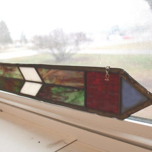 May include: A stained glass arrow with a brown border rests on a windowsill. The arrow features sections of white, red, green, and blue glass. The arrow is approximately 12 inches long.