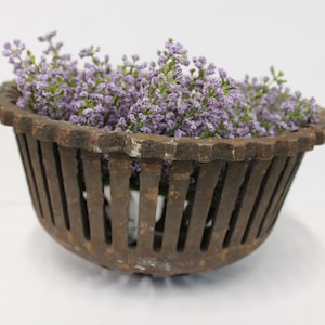 May include: A round, dark brown, weathered cast iron basket with a slatted, open-sided design and a scalloped rim. The basket is filled with a cluster of small, light purple artificial flowers that extend above the top edge of the metal container.