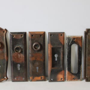 May include: A collection of six antique door hardware pieces, including door plates and escutcheons. The metal hardware has a weathered, aged appearance with shades of brown, copper, and black. Each piece features keyholes and mounting holes.