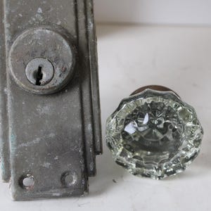 May include: A weathered rectangular metal door plate with a central keyhole cylinder and a separate clear glass doorknob featuring a faceted, multi-sided design. The metal plate has a dull gray finish with visible signs of age and wear.