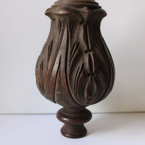 May include: A dark brown, carved wooden furniture leg. The leg features intricate floral and leaf designs, with a bulbous shape and a smaller, rounded base. The wood grain is visible, adding texture to the piece.