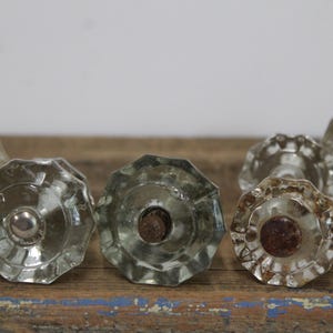 May include: A collection of clear, faceted glass doorknobs. The knobs have a vintage aesthetic, with a central screw and a decorative, octagonal shape. They are arranged on a weathered wooden surface, suggesting a rustic or antique theme.