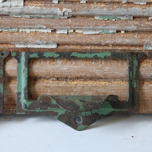 May include: Close-up of a vintage metal label holder with a distressed green finish. The holder is mounted on a weathered wooden surface with peeling paint, showcasing an antique aesthetic. The metal has a rustic, aged appearance.
