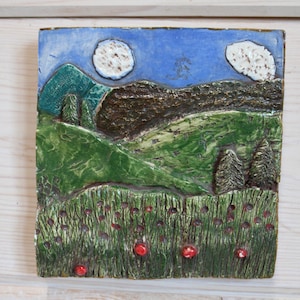 May include: Square ceramic tile with a landscape scene. The artwork features a blue sky with two white, textured moons, green hills, and dark brown mountains. The foreground shows green grass with red accents.