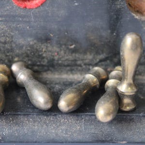 May include: A collection of antique brass drawer pulls and a single, larger handle. The pulls have a rounded top and a cylindrical body. The metal has a tarnished, aged appearance, suggesting vintage hardware.