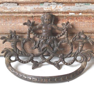 May include: Antique bronze-toned drawer pulls with a cherub and ornate scrollwork design. The pulls have a curved handle and are mounted on a distressed wooden surface, adding a vintage aesthetic.