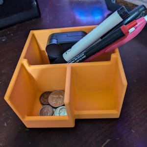 May include: An orange desk organizer with multiple compartments. The organizer holds pens, a small black device, and several coins. The pens are white, black, and pink. The coins are silver and copper-colored.