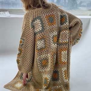 May include: A long, open-front crochet cardigan in a neutral tan color. The back and sleeves feature square patterns in shades of tan, yellow, and green. The cardigan is knee-length and has long sleeves. The overall style is bohemian and handmade.