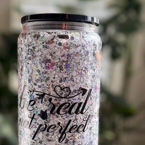 Op de afbeelding: Een doorzichtige glazen beker met een zwart deksel gevuld met zilveren glitter en een zwarte vinyl sticker met de tekst "be real not perfect" met een hart en pijl ontwerp.