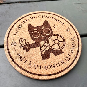 May include: Circular cork coaster featuring a cartoon cat warrior holding a sword and shield. The coaster has text around the edge in French: "GARDEN DU CHAUDRON - PRÊT À AFFRONTER LA CHALEUR". The design is etched into the cork.