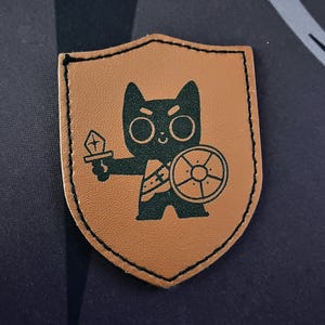 Peut inclure: Écusson en cuir marron en forme de bouclier. Il présente un chat guerrier noir de dessin animé tenant une épée et un bouclier. Le patch a une bordure cousue et est posé sur un fond bleu foncé.
