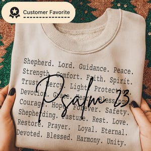 May include: A beige sweatshirt with the text "Psalm 23" in a handwritten font. The sweatshirt features a list of words like "Shepherd," "Faith," and "Blessings" in a black, sans-serif font. The words are arranged in columns.