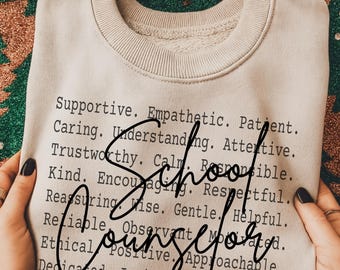 May include: A beige sweatshirt with the words "School Counselor" in a cursive font. The sweatshirt is printed with a list of positive attributes such as "Supportive," "Empathetic," and "Kind." The background is a festive pattern.