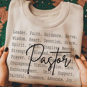 May include: A beige sweatshirt with the word "Pastor" in a cursive font. The shirt is printed with words like "Leader," "Faith," and "Courageous." The sweatshirt is folded and held by a person.