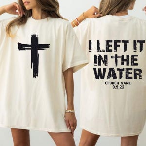Pode incluir: Camiseta creme com uma grande cruz preta na frente. A parte de trás da camiseta apresenta o texto "I LEFT IT IN THE WATER" em preto, com "CHURCH NAME" e a data "9,9,22" abaixo.