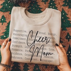 Op de afbeelding: Een beige sweatshirt met de woorden "Cheer Mom" in een cursief lettertype. Het sweatshirt heeft een lijst met woorden als "Supportive", "Encouraging" en "Confidence" gedrukt in een zwarte, schreefloze letter.