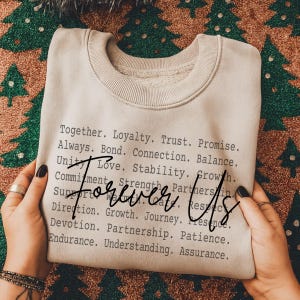 Può includere: Una felpa beige con le parole "Forever Us" in corsivo. La felpa è stampata con un elenco di parole tra cui "Insieme", "Lealtà" e "Fiducia". La felpa è piegata e tenuta in mano.