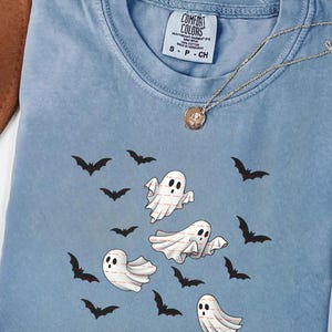 May include: A light blue Comfort Colors t-shirt featuring a Halloween design. The shirt has a graphic of white ghosts with black eyes and bats. A gold necklace with a pendant is also visible.