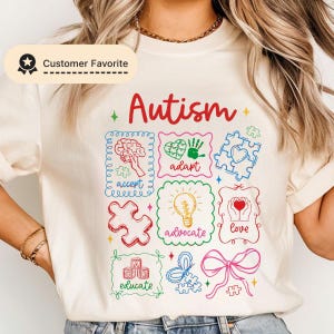 May include: Cream-colored t-shirt with the word "Autism" in red script. The shirt features colorful graphics including puzzle pieces, a brain, a heart, and the words "accept," "adapt," "advocate," "love," and "educate."