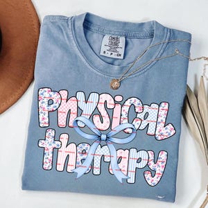May include: A blue Comfort Colors t-shirt with the words "Physical Therapy" in a floral and checkered design, accented with a light blue bow. A gold necklace is draped across the shirt.