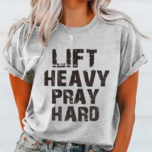May include: A light gray t-shirt with the words "LIFT HEAVY PRAY HARD" printed in a distressed black font. The shirt has short sleeves and a crew neck. The model is wearing blue jeans.