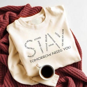 Op de afbeelding: Crèmekleurige sweatshirt met het woord "SAY" gevormd door positieve woorden als "geliefd" en "dapper". De tekst "TOMORROW NEEDS YOU" staat eronder. Een kopje koffie staat onderaan. De sweatshirt ligt op een bordeauxrode gebreide deken.