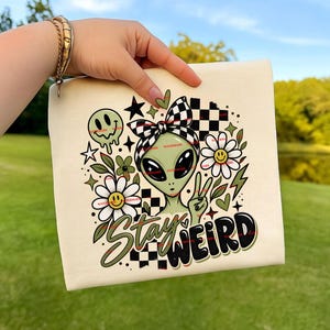 Pode incluir: Uma bolsa de lona cor de creme com uma estampa gráfica de um alienígena verde usando uma faixa de cabeça xadrez preta e branca. O design inclui margaridas com rostos sorridentes, um smiley derretendo, estrelas e o texto Stay Weird em preto.
