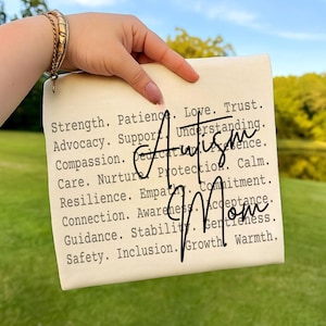 Puede incluir: Un letrero rectangular color crema con las palabras "Autism Mom" en una fuente manuscrita. El letrero enumera palabras como "Fuerza", "Paciencia" y "Amor" en una fuente de máquina de escribir. El fondo es una escena borrosa al aire libre.