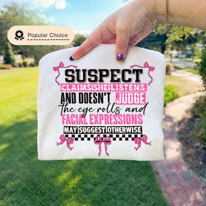 Puede incluir: Una camiseta blanca con el texto "SUSPECT CLAIMSISHEILISTENS AND DOESN'T JUDGE the eye rolls and FACIAL EXPRESSIONS MAY SUGGEST OTHERWISE" en negro y rosa. La camiseta tiene lazos rosas y una insignia de Popular Choice.