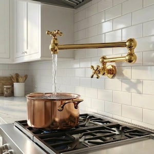 Op de afbeelding: Een koperen pan op een gasfornuis wordt gevuld met water uit een goudkleurige, aan de muur gemonteerde kraan. De kraan heeft kruisvormige handgrepen en een zwenkarm. De keuken heeft witte kasten en een witte tegelachterwand.