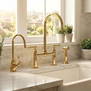 May include: A gold-toned kitchen faucet set with a bridge design, featuring a high-arc spout and separate side sprayer. The faucet is installed on a white countertop above a white farmhouse sink. Small potted plants sit on the windowsill.