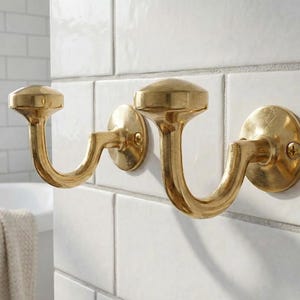 Puede incluir: Dos perchas de pared doradas con un diseño curvo. Cada gancho tiene una base circular y una parte superior decorativa. Los ganchos están montados sobre baldosas cuadradas blancas, lo que sugiere un baño o similar.