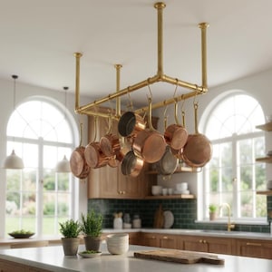 Puede incluir: Una cocina con un estante para ollas de latón suspendido del techo, que sostiene varias ollas de cobre. La cocina cuenta con gabinetes de madera, una encimera blanca y grandes ventanas arqueadas, creando un ambiente luminoso. Hay plantas en macetas y una tabla de cortar en la encimera.