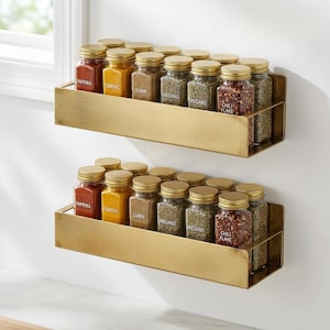Peut inclure: Deux étagères à épices en laiton fixées au mur blanc, chacune contenant des pots à épices en verre avec des couvercles dorés. Les épices comprennent du paprika, du curcuma, du cumin, de l'origan et des flocons de piment. Les pots sont rangés, pour la cuisine.