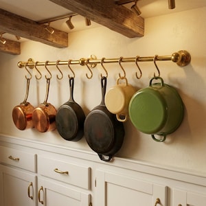 Könnte beinhalten: Ein Messing-Topfgestell mit verschiedenen Töpfen und Pfannen, die daran hängen. Die Sammlung umfasst Kupferpfannen, gusseiserne Bratpfannen und einen grünen Emailletopf. Das Gestell ist über weißen Schränken montiert.