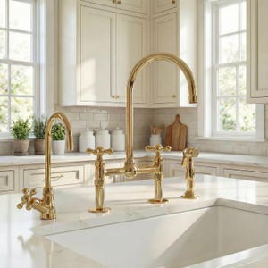 May include: A gold kitchen tap set with a bridge design, featuring a high-arc spout and cross-handle detailing. The set includes a side sprayer. The tap is installed on a white countertop in a bright kitchen setting.